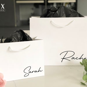 May include: Two white gift bags with black tissue paper inside. The larger bag has the name "Rachel" written on it in black script. The smaller bag has the name "Sarah" written on it in black script.