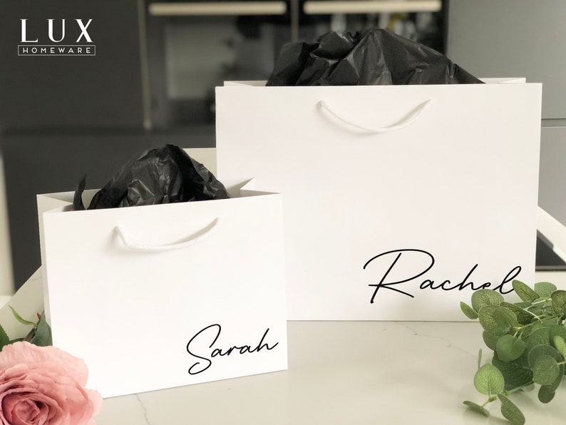 May include: Two white gift bags with black tissue paper inside. The larger bag has the name "Rachel" written on it in black script. The smaller bag has the name "Sarah" written on it in black script.