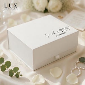 May include: A white gift box with a lid, featuring the names "Sarah & Matt" and the date "07.09.2026" in elegant script. The box has a satin ribbon pull and is surrounded by floral accents and pearls.