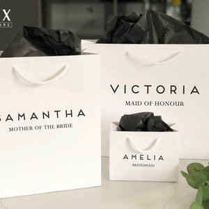 May include: Three white gift bags with black tissue paper inside. The bags are labeled with the names "Samantha, Mother of the Bride", "Victoria, Maid of Honour", and "Amelia, Bridesmaid".
