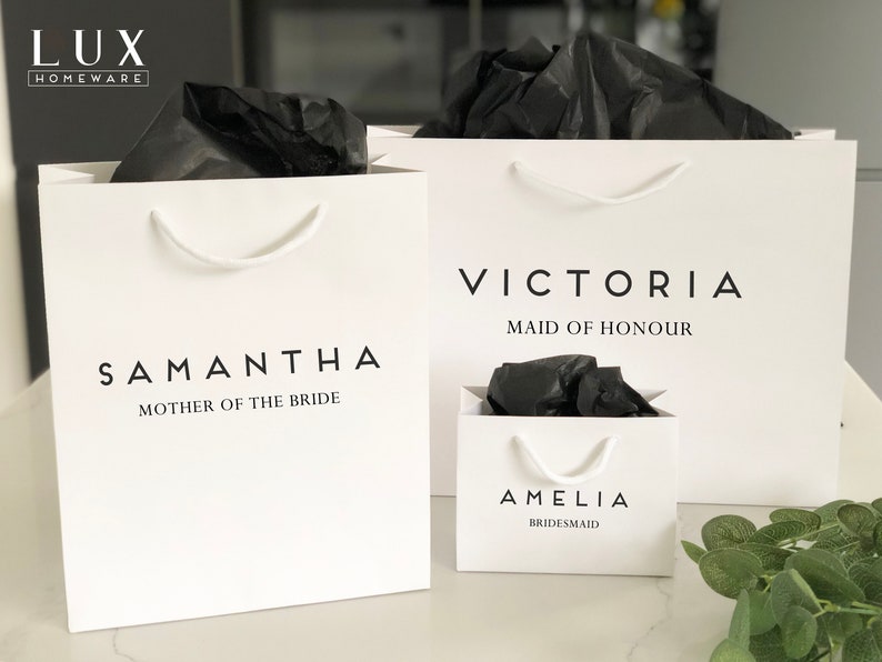 May include: Three white gift bags with black tissue paper inside. The bags are labeled with the names "Samantha, Mother of the Bride", "Victoria, Maid of Honour", and "Amelia, Bridesmaid".