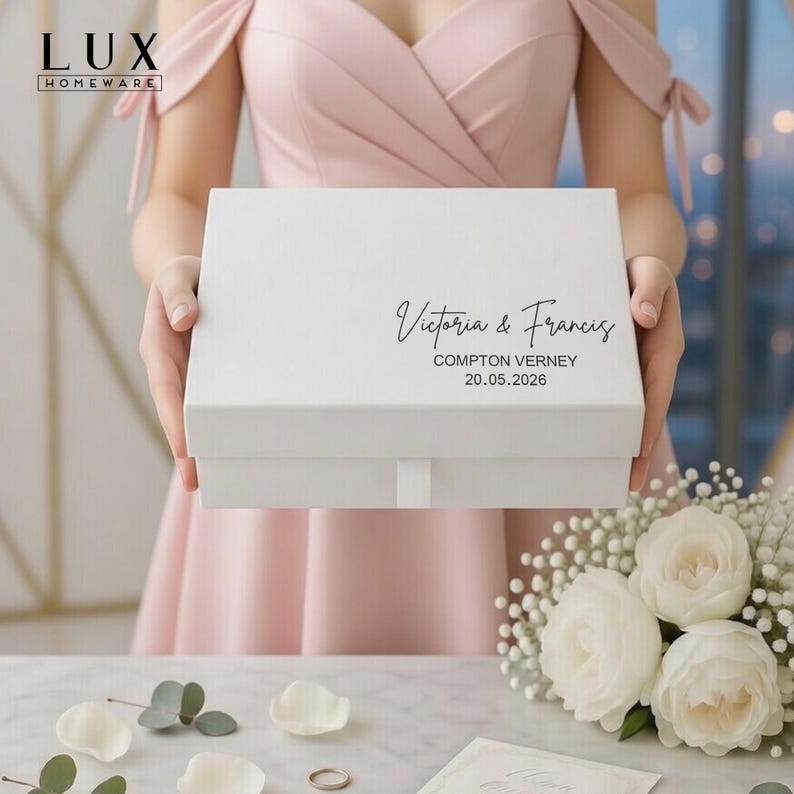 May include: A white gift box bearing the names "Victoria & Francis" and the date "20.05.2026" in black script. The box is held by a person wearing a pink dress. The LUX HOMEWARE logo is in the top left corner.