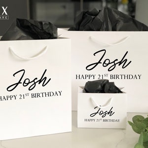 May include: Three white gift bags with black handles and black tissue paper inside. The bags are personalized with the name "Josh" and the text "Happy 21st Birthday".
