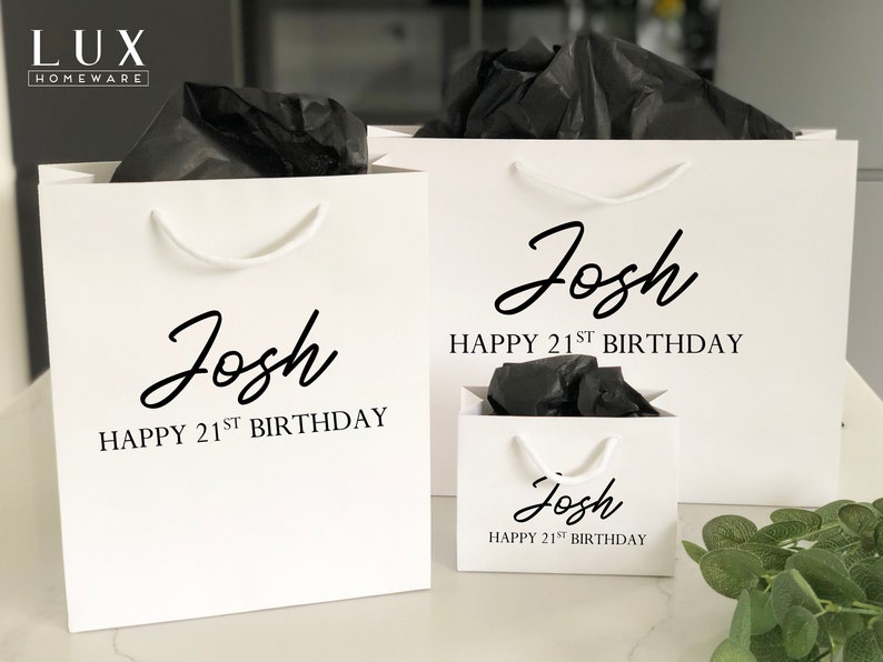May include: Three white gift bags with black handles and black tissue paper inside. The bags are personalized with the name "Josh" and the text "Happy 21st Birthday".