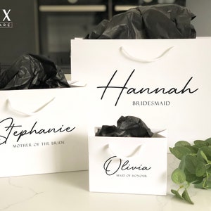 May include: Three white gift bags with black handles and black tissue paper inside. The bags are personalised with the names "Hannah Bridesmaid", "Stephanie Mother of the Bride", and "Olivia Maid of Honour".