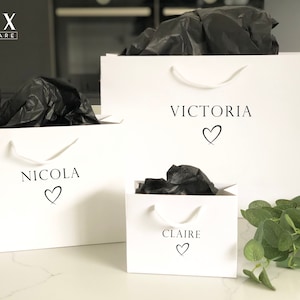 May include: Three white gift bags with black handles and black tissue paper inside. The bags are labeled with the names "Victoria", "Nicola", and "Claire" with a heart symbol next to each name.