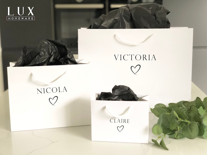May include: Three white gift bags with black handles and black tissue paper inside. The bags are labeled with the names "Victoria", "Nicola", and "Claire" with a heart symbol next to each name.