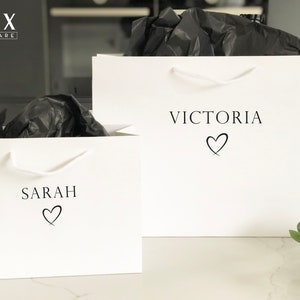May include: Two white gift bags with black handles. The bag on the left has the name "SARAH" printed on it with a heart symbol. The bag on the right has the name "VICTORIA" printed on it with a heart symbol.