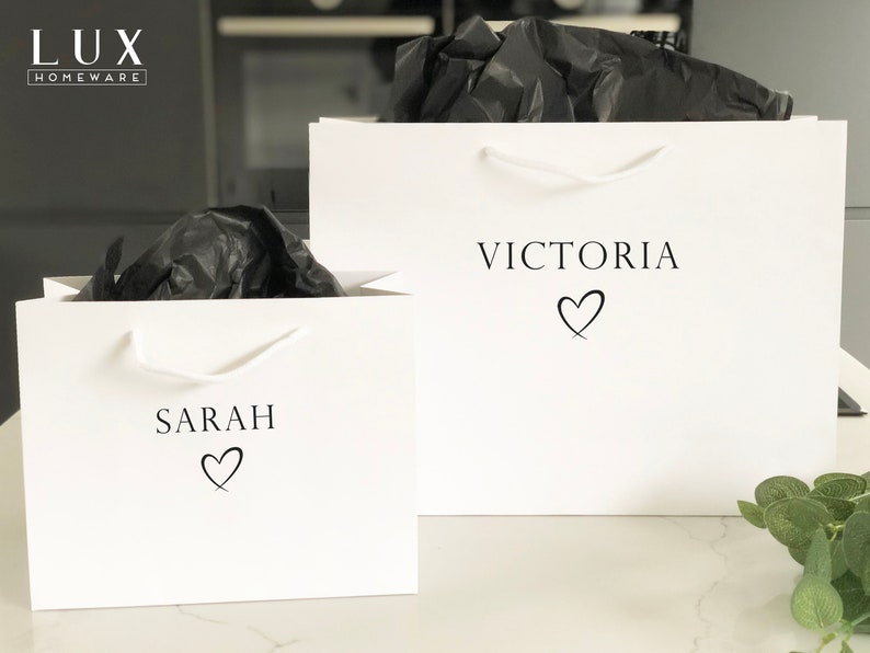 May include: Two white gift bags with black handles. The bag on the left has the name "SARAH" printed on it with a heart symbol. The bag on the right has the name "VICTORIA" printed on it with a heart symbol.