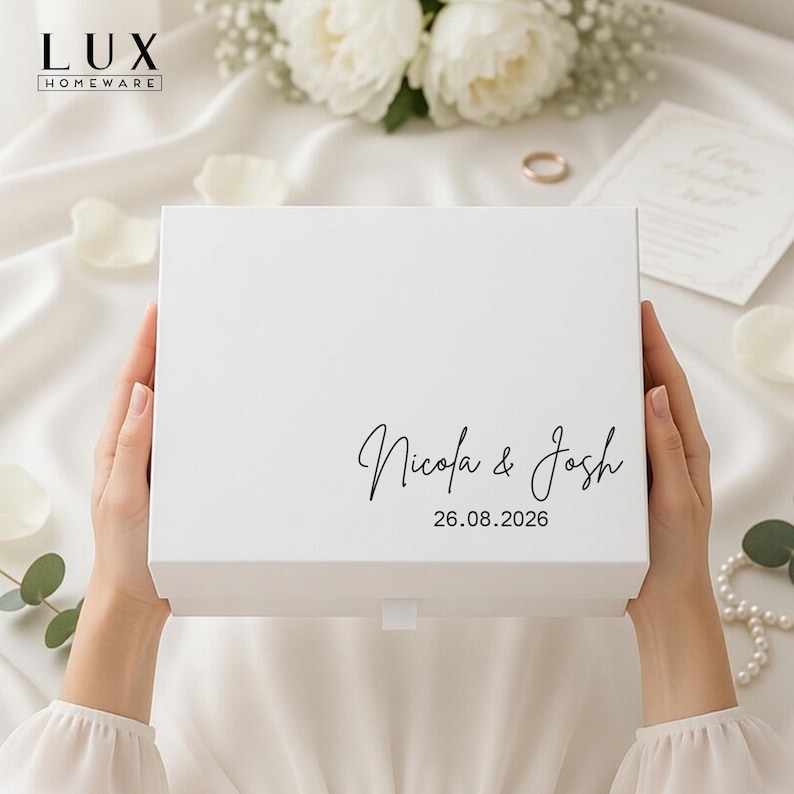 May include: A white gift box, personalised with the names "Nicola & Josh" and the date "26.08.2026" in a stylish script. The box is held by hands, with a backdrop of white fabric, flowers, and wedding accessories.