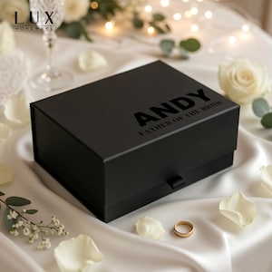 K&ouml;nnte beinhalten: Eine schwarze Geschenkbox mit dem Namen "ANDY" und dem Text "FATHER OF THE BRIDE" auf dem Deckel. Die rechteckige Box liegt auf einer wei&szlig;en Satin-Oberfl&auml;che, umgeben von wei&szlig;en Rosenbl&auml;ttern und einem goldenen Ehering.