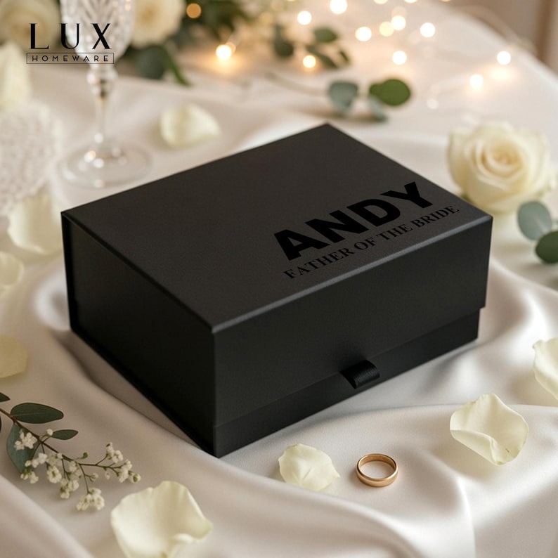 K&ouml;nnte beinhalten: Eine schwarze Geschenkbox mit dem Namen "ANDY" und dem Text "FATHER OF THE BRIDE" auf dem Deckel. Die rechteckige Box liegt auf einer wei&szlig;en Satin-Oberfl&auml;che, umgeben von wei&szlig;en Rosenbl&auml;ttern und einem goldenen Ehering.