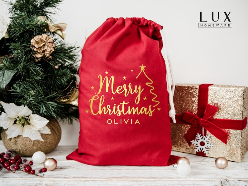 Pode incluir: Uma sacola de presente de Natal vermelha com fecho de cord&atilde;o. A sacola apresenta texto dourado que diz "Merry Christmas" e um desenho de &aacute;rvore de Natal. Uma caixa de presente dourada com lantejoulas e uma fita vermelha est&aacute; no fundo.