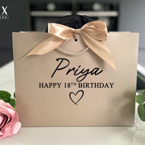 K&ouml;nnte beinhalten: Eine beige Geschenkverpackung mit einer Satinschleife und Seilgriffen. Die Tasche ist mit dem Namen "Priya", dem Text "HAPPY 18TH BIRTHDAY" und einem Herzsymbol personalisiert. Eine rosa Rose und Gr&uuml;nzeug sind im Vordergrund.