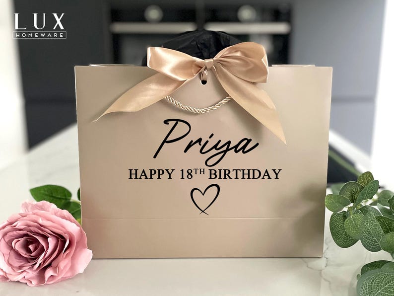 K&ouml;nnte beinhalten: Eine beige Geschenkverpackung mit einer Satinschleife und Seilgriffen. Die Tasche ist mit dem Namen "Priya", dem Text "HAPPY 18TH BIRTHDAY" und einem Herzsymbol personalisiert. Eine rosa Rose und Gr&uuml;nzeug sind im Vordergrund.