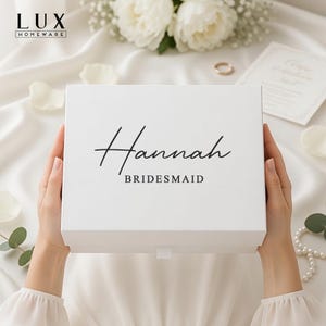 May include: A white gift box, featuring the name "Hannah" and the word "Bridesmaid" in black script. The box is held by a person, with a white background and floral accents.