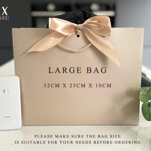 K&ouml;nnte beinhalten: Eine gro&szlig;e beige Geschenkt&uuml;te mit cremefarbener Schleife und Kordelgriffen. Auf der T&uuml;te steht "LARGE BAG" und sie misst 32 cm x 25 cm x 10 cm. Neben der T&uuml;te liegt ein silbernes Smartphone. Von LUX HOMEWARE.