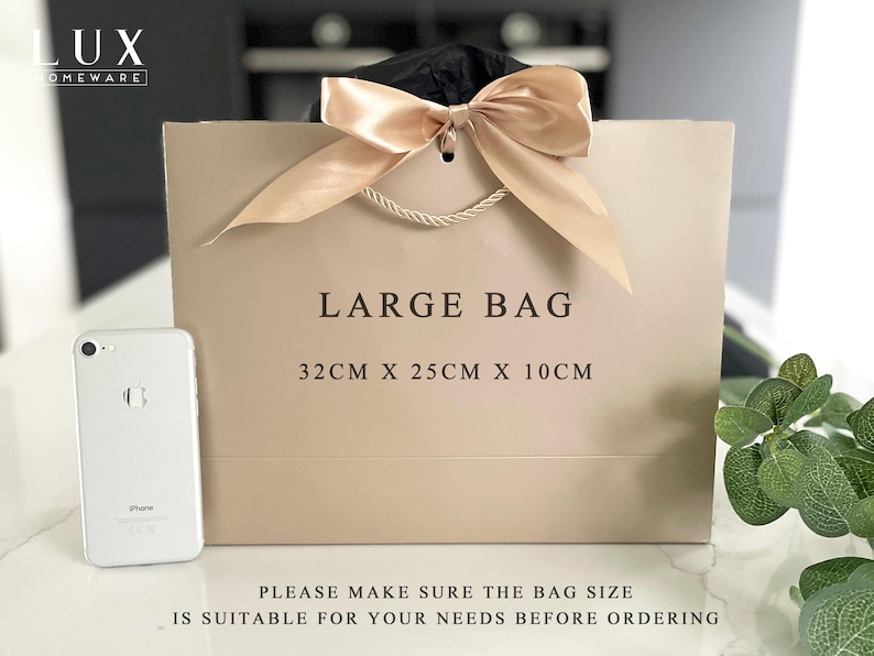 K&ouml;nnte beinhalten: Eine gro&szlig;e beige Geschenkt&uuml;te mit cremefarbener Schleife und Kordelgriffen. Auf der T&uuml;te steht "LARGE BAG" und sie misst 32 cm x 25 cm x 10 cm. Neben der T&uuml;te liegt ein silbernes Smartphone. Von LUX HOMEWARE.