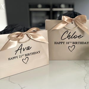 K&ouml;nnte beinhalten: Zwei beige Geschenkt&uuml;ten mit goldenen Satinb&auml;ndern. Auf einer T&uuml;te steht "Ava HAPPY 21ST BIRTHAY" und auf der anderen "Chloe HAPPY 18TH BIRTHDAY" in schwarzer Schrift. Jede T&uuml;te hat ein Herzsymbol unter dem Text. Die T&uuml;ten stehen auf einer wei&szlig;en Oberfl&auml;che.