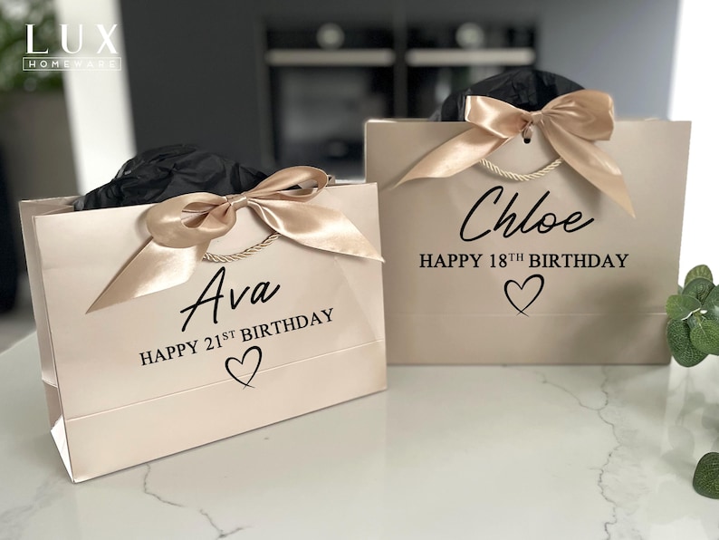 K&ouml;nnte beinhalten: Zwei beige Geschenkt&uuml;ten mit goldenen Satinb&auml;ndern. Auf einer T&uuml;te steht "Ava HAPPY 21ST BIRTHAY" und auf der anderen "Chloe HAPPY 18TH BIRTHDAY" in schwarzer Schrift. Jede T&uuml;te hat ein Herzsymbol unter dem Text. Die T&uuml;ten stehen auf einer wei&szlig;en Oberfl&auml;che.