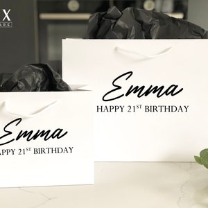 May include: Two white gift bags with white handles. The bags are personalized with the name "Emma" and the text "Happy 21st Birthday".