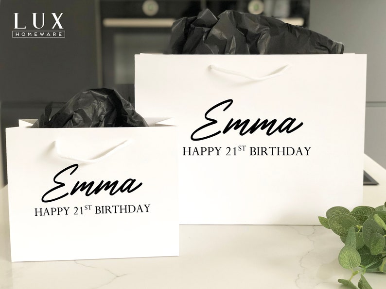 May include: Two white gift bags with white handles. The bags are personalized with the name "Emma" and the text "Happy 21st Birthday".