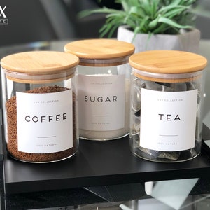 May include: Three glass jars with bamboo lids and labels that say "Coffee", "Sugar", and "Tea". The jars are sitting on a black tray.