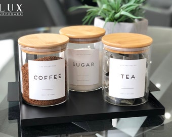 May include: Three glass jars with bamboo lids and labels that say "Coffee", "Sugar", and "Tea". The jars are sitting on a black tray.