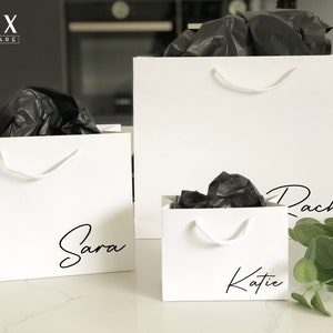 May include: Three white gift bags with black tissue paper inside. The bags have handles and are personalised with the names "Sara", "Katie", and "Rachel" in black script.