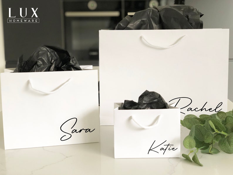 May include: Three white gift bags with black tissue paper inside. The bags have handles and are personalised with the names "Sara", "Katie", and "Rachel" in black script.