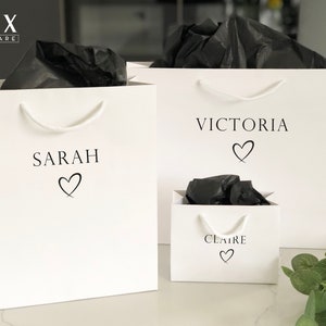 May include: Three white gift bags with black tissue paper inside. The bags are personalized with the names "Sarah", "Victoria", and "Claire" and a heart symbol.