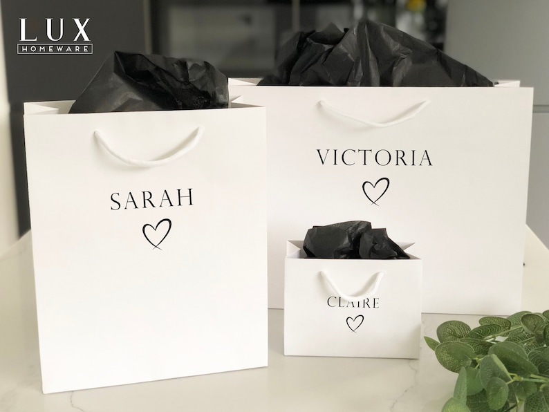 May include: Three white gift bags with black tissue paper inside. The bags are personalized with the names "Sarah", "Victoria", and "Claire" and a heart symbol.