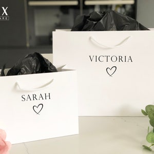 May include: Two white gift bags with black tissue paper inside. The bags have black text that says "Sarah" and "Victoria" with a heart symbol below each name.