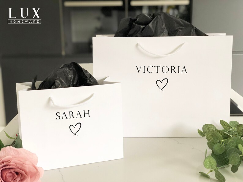 May include: Two white gift bags with black tissue paper inside. The bags have black text that says "Sarah" and "Victoria" with a heart symbol below each name.