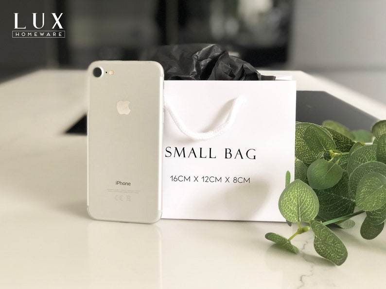 May include: A white gift bag with black text that reads "SMALL BAG 16CM X 12CM X 8CM". A silver iPhone is in the foreground.