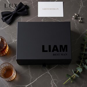 K&ouml;nnte beinhalten: Eine schwarze Geschenkbox mit dem Namen "LIAM" und dem Text "BEST MAN" aufgedruckt. Eine schwarze Fliege, eine Karte mit der Aufschrift "GROOMSMAN" und ein Paar Manschettenkn&ouml;pfe sind ebenfalls sichtbar. Zwei Gl&auml;ser mit bernsteinfarbener Fl&uuml;ssigkeit befinden sich links.