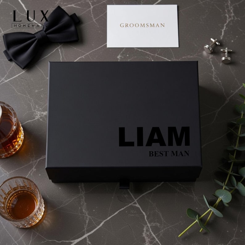 K&ouml;nnte beinhalten: Eine schwarze Geschenkbox mit dem Namen "LIAM" und dem Text "BEST MAN" aufgedruckt. Eine schwarze Fliege, eine Karte mit der Aufschrift "GROOMSMAN" und ein Paar Manschettenkn&ouml;pfe sind ebenfalls sichtbar. Zwei Gl&auml;ser mit bernsteinfarbener Fl&uuml;ssigkeit befinden sich links.