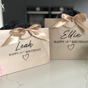 K&ouml;nnte beinhalten: Zwei beige Geschenkt&uuml;ten mit Satinb&auml;ndern und Kordelgriffen. Eine T&uuml;te tr&auml;gt die Aufschrift "Leah HAPPY 21ST BIRTHDAY" mit einem Herz, die andere "Ellie HAPPY 18TH BIRTHDAY" mit einem Herz. Die T&uuml;ten stehen auf einer wei&szlig;en Oberfl&auml;che.