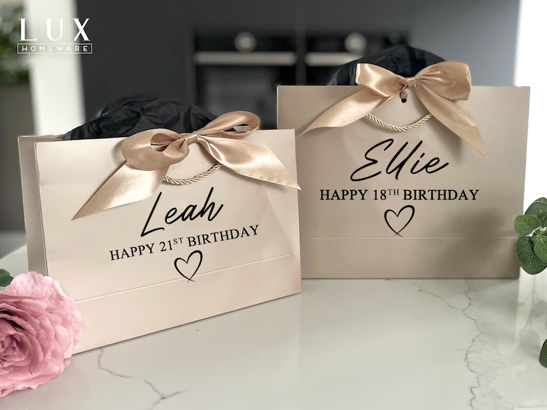 K&ouml;nnte beinhalten: Zwei beige Geschenkt&uuml;ten mit Satinb&auml;ndern und Kordelgriffen. Eine T&uuml;te tr&auml;gt die Aufschrift "Leah HAPPY 21ST BIRTHDAY" mit einem Herz, die andere "Ellie HAPPY 18TH BIRTHDAY" mit einem Herz. Die T&uuml;ten stehen auf einer wei&szlig;en Oberfl&auml;che.