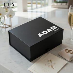 K&ouml;nnte beinhalten: Eine schwarze Geschenkbox mit dem Namen "ADAM" und dem Wort "GROOM" in Wei&szlig; aufgedruckt. Die Box steht auf einer Marmoroberfl&auml;che, mit Eheringen, einem Sektglas und einer Hochzeitseinladung in der N&auml;he. Die Box hat einen Magnetverschluss.
