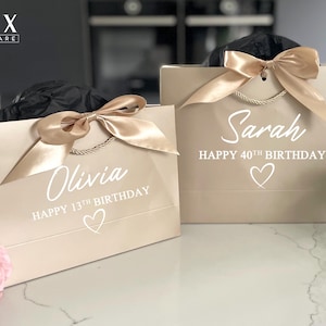 K&ouml;nnte beinhalten: Zwei beige Geschenkt&uuml;ten mit goldenen Satinb&auml;ndern. Auf einer T&uuml;te steht "Olivia HAPPY 13TH BIRTHDAY" und auf der anderen "Sarah HAPPY 40TH BIRTHDAY". Beide T&uuml;ten haben ein Herzsymbol. Die T&uuml;ten haben goldene Kordelgriffe.