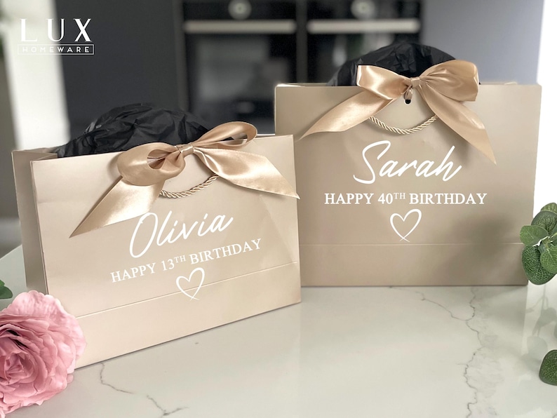 K&ouml;nnte beinhalten: Zwei beige Geschenkt&uuml;ten mit goldenen Satinb&auml;ndern. Auf einer T&uuml;te steht "Olivia HAPPY 13TH BIRTHDAY" und auf der anderen "Sarah HAPPY 40TH BIRTHDAY". Beide T&uuml;ten haben ein Herzsymbol. Die T&uuml;ten haben goldene Kordelgriffe.