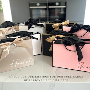 K&ouml;nnte beinhalten: Eine Auswahl an personalisierten Geschenkbeuteln in Beige, Schwarz und Rosa, jeweils mit einer Satinschleife und einem Namen in Schreibschrift. Die Beutel sind auf einer wei&szlig;en Arbeitsplatte angeordnet. Der Text auf dem Bild lautet: BESUCHEN SIE UNSERE LISTINGS F&Uuml;R UNSERE GESAMTE AUSWAHL AN PERSONALISIERTEN GESCHENKTASCHEN.