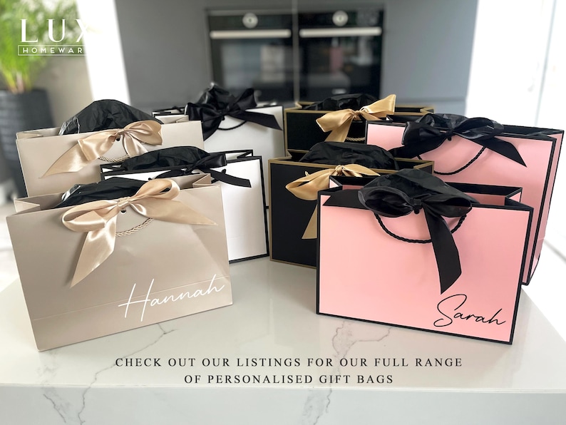 K&ouml;nnte beinhalten: Eine Auswahl an personalisierten Geschenkbeuteln in Beige, Schwarz und Rosa, jeweils mit einer Satinschleife und einem Namen in Schreibschrift. Die Beutel sind auf einer wei&szlig;en Arbeitsplatte angeordnet. Der Text auf dem Bild lautet: BESUCHEN SIE UNSERE LISTINGS F&Uuml;R UNSERE GESAMTE AUSWAHL AN PERSONALISIERTEN GESCHENKTASCHEN.