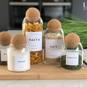May include: Five glass jars with cork lids and labels. The jars contain pasta, oats, sugar, parsley, and basil. The labels are white with black text and say "Pasta", "Oats", "Sugar", "Parsley", and "Basil". The jars are on a wooden chopping board.