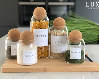 Könnte beinhalten: Fünf Glasbehälter mit Korkdeckeln und Etiketten. Die Behälter enthalten Pasta, Haferflocken, Zucker, Petersilie und Basilikum. Die Etiketten sind weiß mit schwarzem Text und lauten "Pasta", "Haferflocken", "Zucker", "Petersilie" und "Basilikum". Die Behälter stehen auf einem Holzbrett.