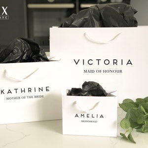 May include: Three white gift bags with black tissue paper inside. The bags are labeled with the names "Amelia Bridesmaid", "Victoria Maid of Honour", and "Kathrine Mother of the Bride".