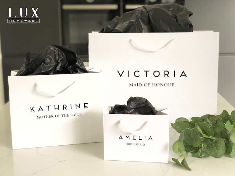 May include: Three white gift bags with black tissue paper inside. The bags are labeled with the names "Amelia Bridesmaid", "Victoria Maid of Honour", and "Kathrine Mother of the Bride".