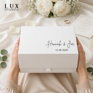 May include: A white gift box, featuring the names "Hannah & Joe" and the date "12.06.2026" in black script. The box is held by hands, set against a backdrop of white fabric and floral arrangements.