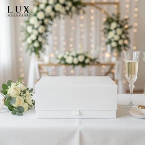 May include: A white gift box with a ribbon pull on a white tablecloth. A champagne flute, wedding rings, and a bouquet of white roses are also on the table. The background features a floral arch and string lights.